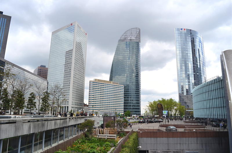 12 - La Défense business district in Paris, France, featuring prominent skyscrapers like the curved Tour First (formerly Tour Assur) and Tour Égée under a cloudy sky